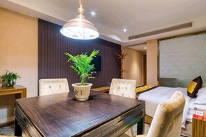Une chambre d’hôtel avec une table à manger en bois, quatre chaises, une plante en pot, un lit avec une tête de lit blanche et une grande fenêtre.