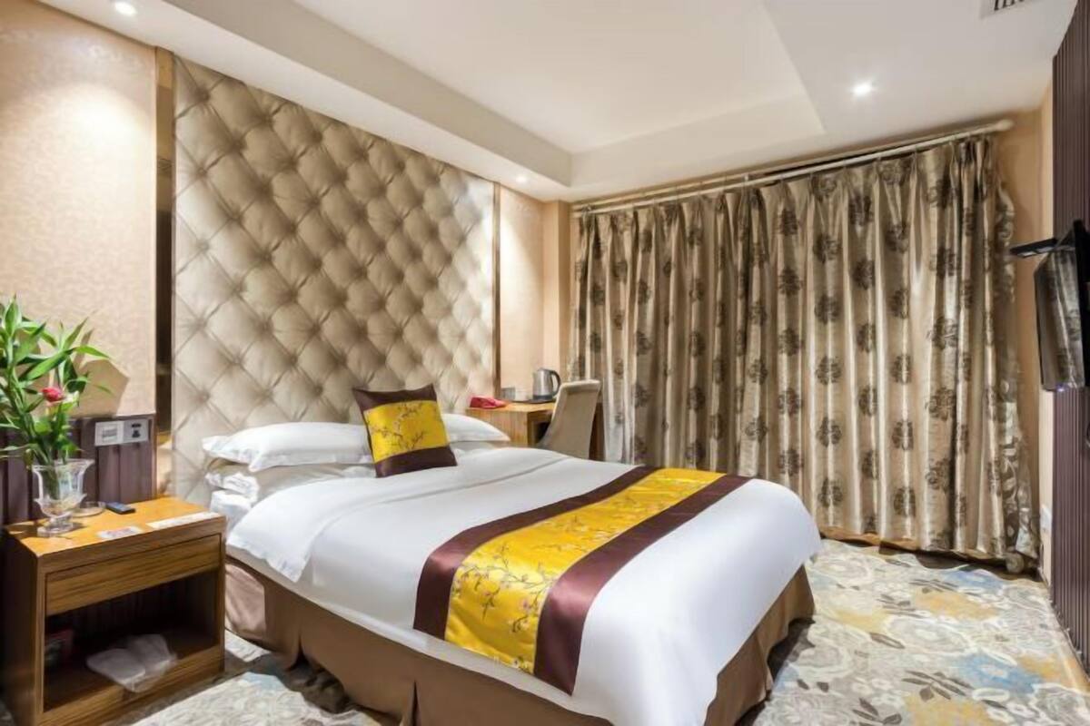 Una camera d'hotel con una testiera trapuntata, un letto con biancheria gialla e bianca, un comodino in legno, una scrivania con una sedia e una finestra con tende floreali.