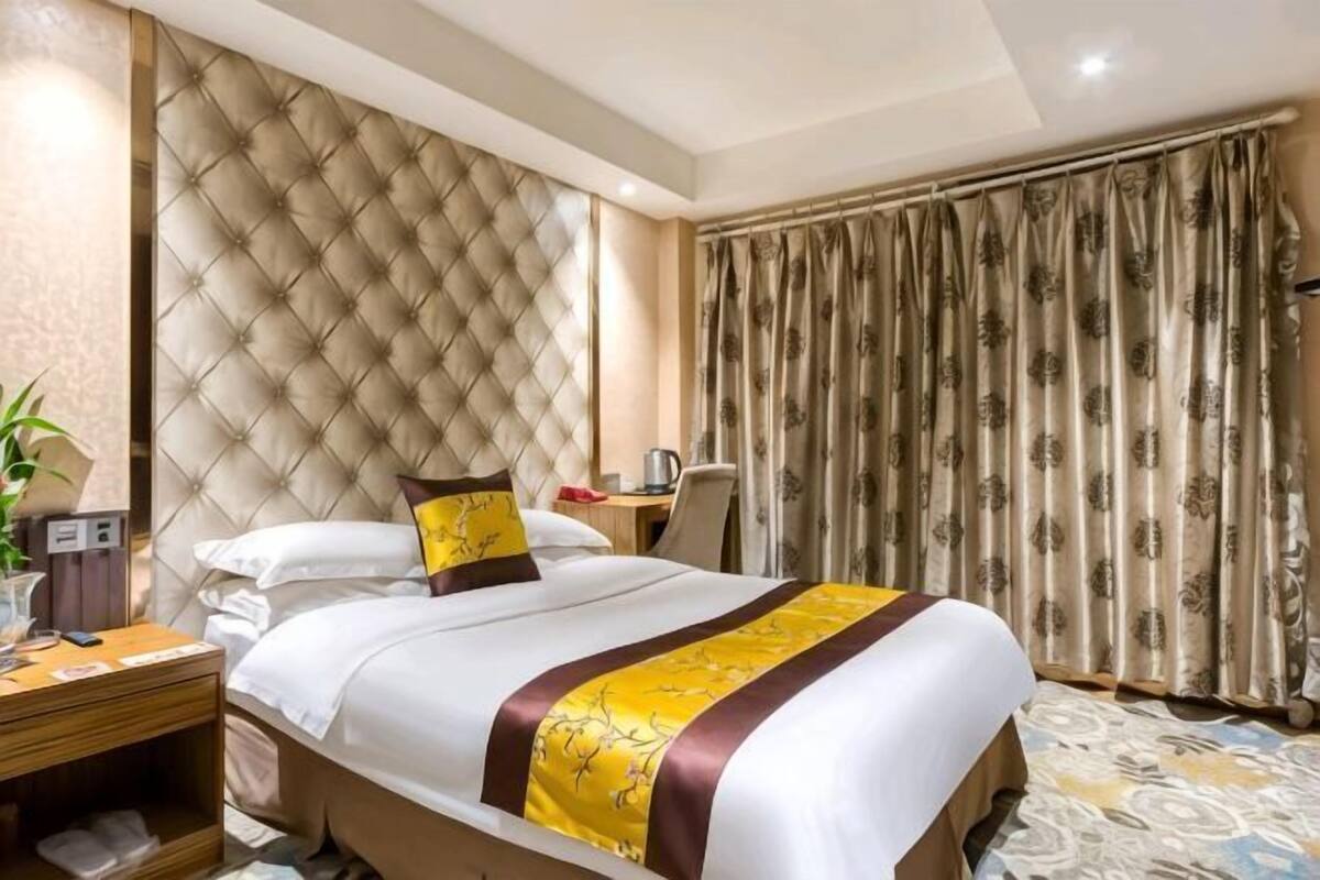 Una camera d'albergo con una testata imbottita, un letto con copriletto giallo e marrone, un comodino con una lampada e un'ampia finestra con tende.