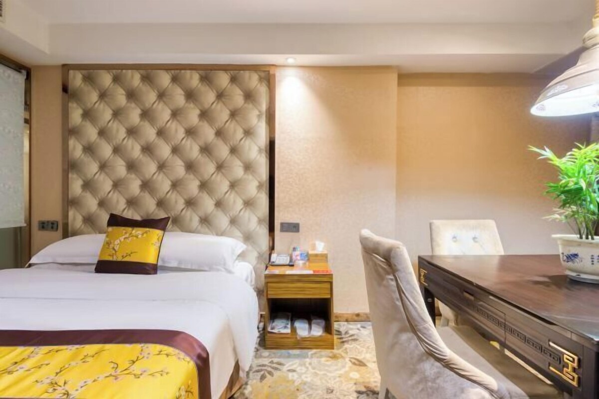 A hotel room with a large tufted headboard, a bed with white linens, a wooden nightstand with a lamp, a dining table with chairs, and a potted plant.