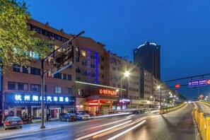 Een verlichte stadstraat 's nachts met gebouwen, een heldere hemel en een weg met lichtsporen van voorbijrijdende voertuigen.