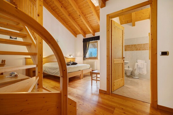 Un intérieur en bois comprenant un lit, une petite table, un radiateur et une salle de bain visible à travers une porte ouverte.