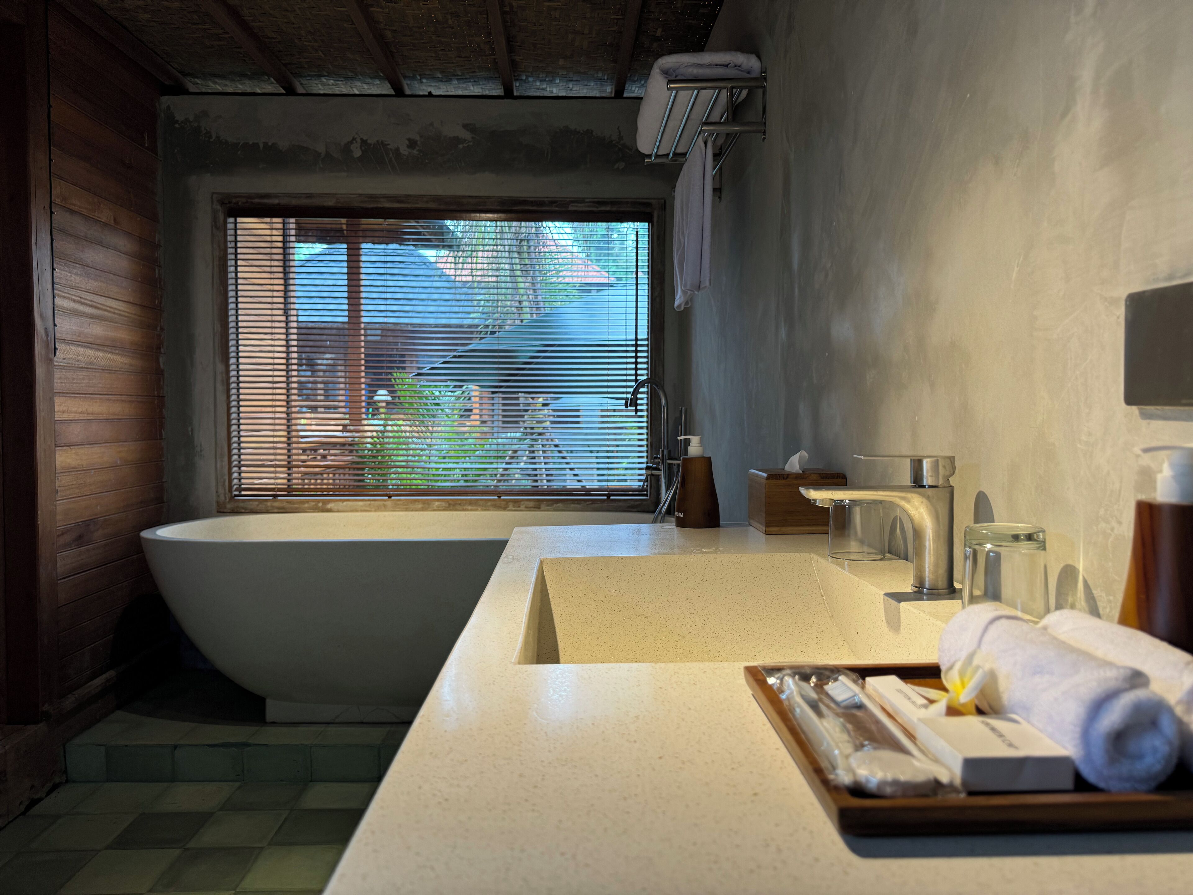 A modern bathroom with a freestanding bathtub, a large window with blinds, a countertop with toiletries, and a towel rack.