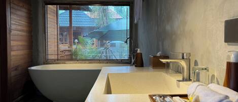 A modern bathroom with a freestanding bathtub, a large window with blinds, a countertop with toiletries, and a towel rack.