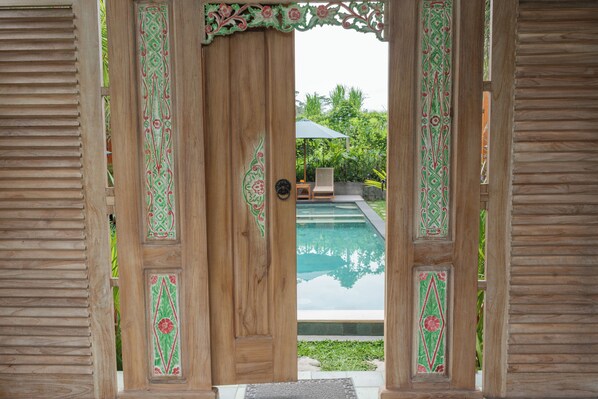 Une porte en bois ornée de motifs complexes, donnant sur un espace piscine équipé d’une chaise et d’un parasol.