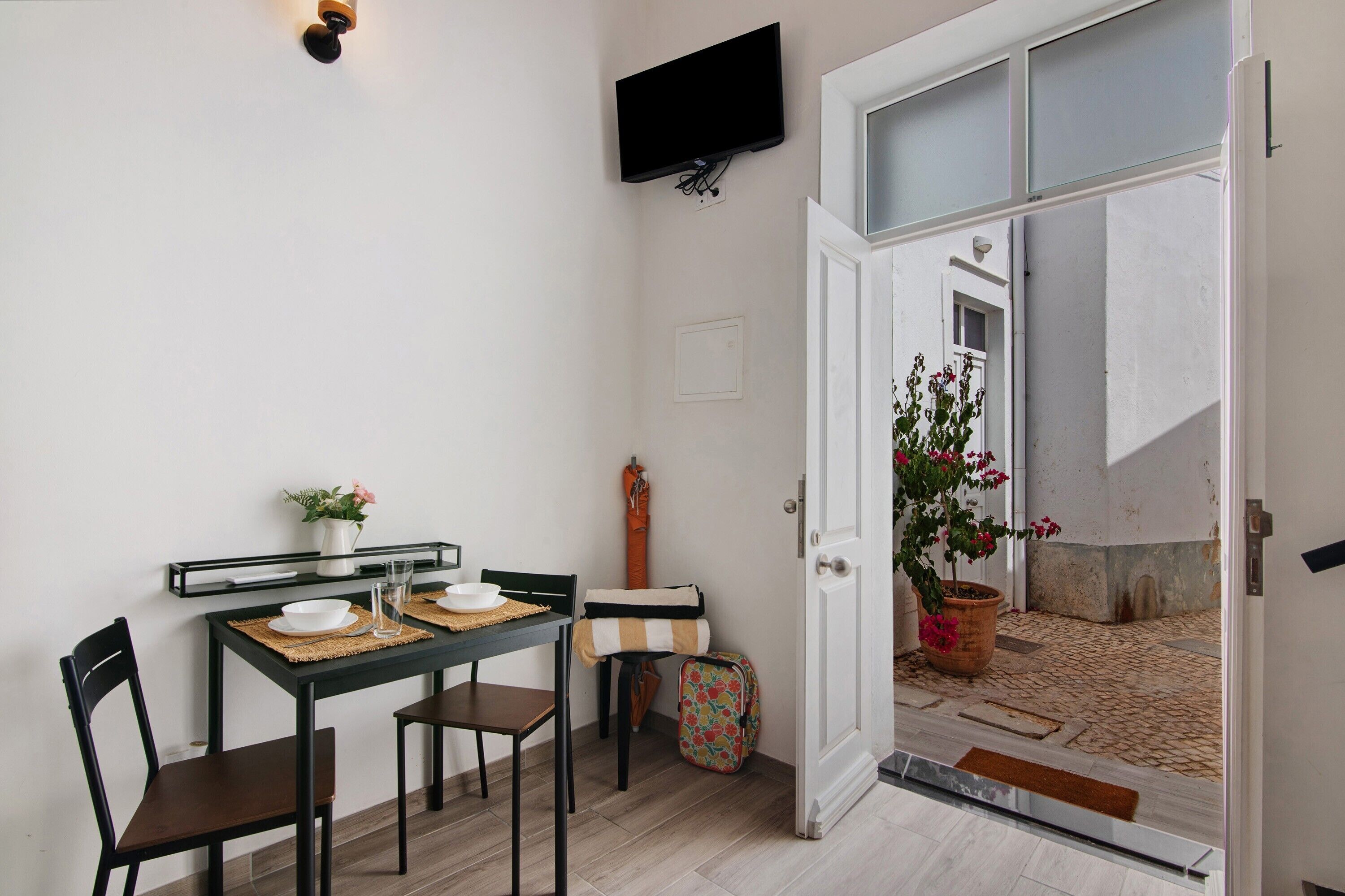 Un comedor con una mesa, sillas, un televisor montado en la pared y una puerta que da a un espacio exterior con una planta en maceta.
