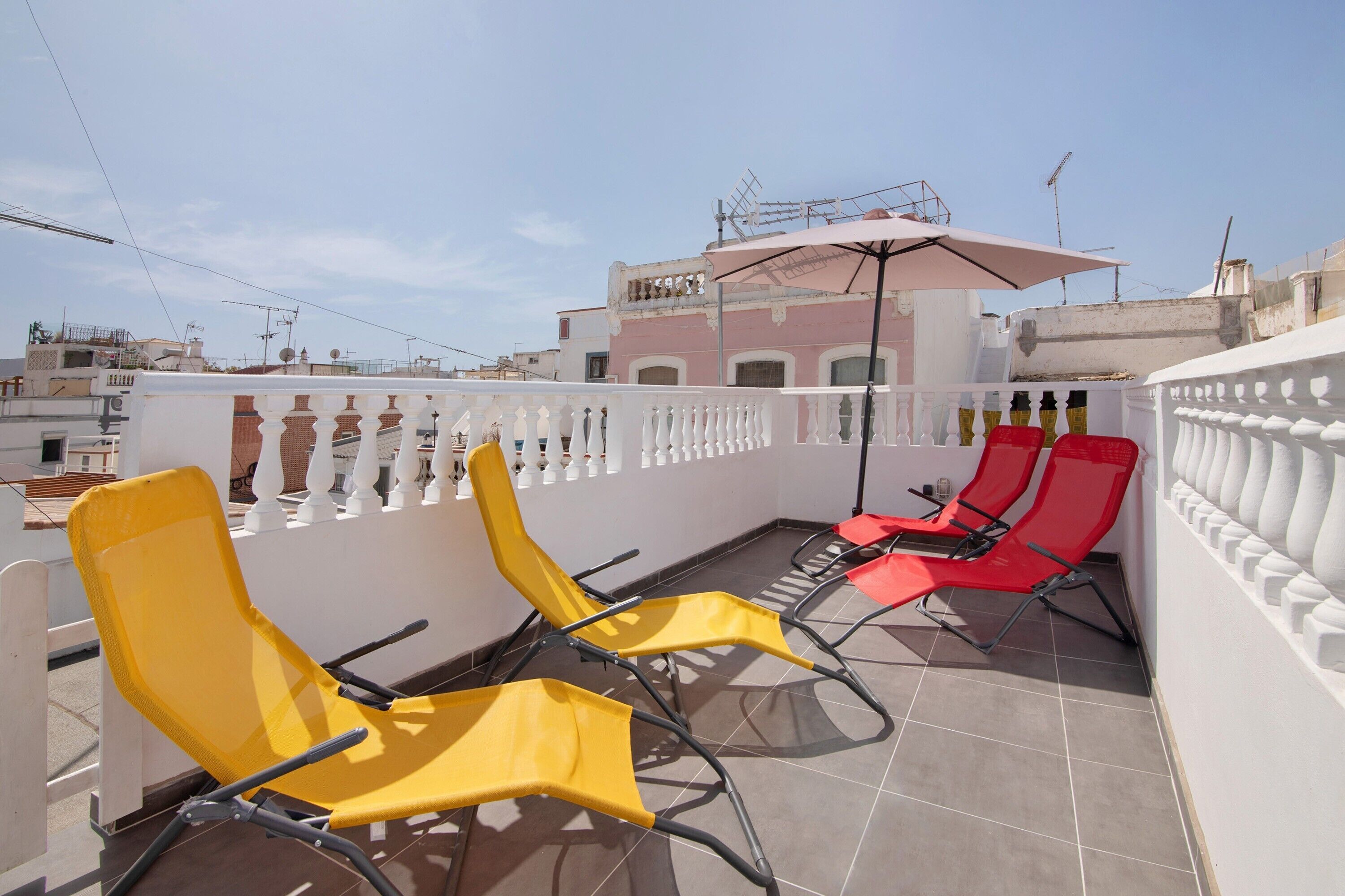Una terraza en la azotea con sillones reclinables amarillos y rojos, barandilla blanca y vista despejada del paisaje urbano.