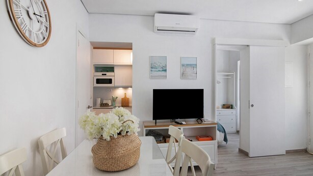 Een moderne keuken met witte kasten, een flatscreen televisie en een eethoek met een witte tafel en stoelen.
