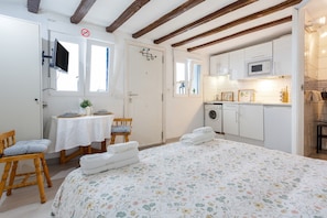 Un dormitorio con cama, una pequeña mesa con sillas, televisor y una zona de cocina con armarios y electrodomésticos.