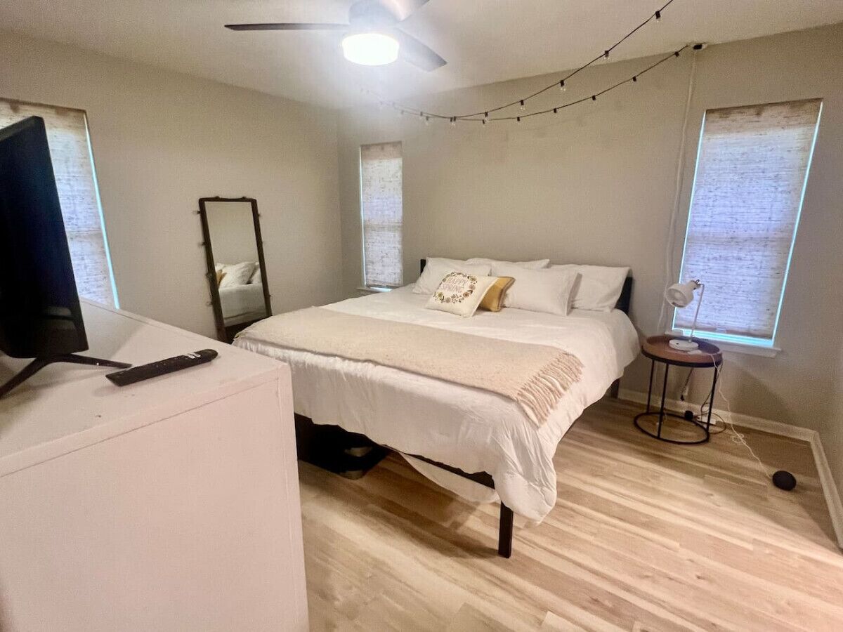 Une chambre à coucher avec un lit, un bureau, une chaise, un miroir et une fenêtre avec des rideaux.