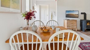 Un comedor con una mesa de madera, sillas blancas, un jarrón de flores y un televisor sobre un mueble de madera.