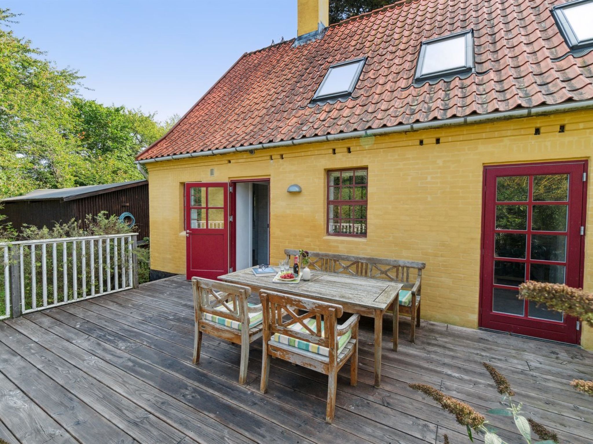 A wooden deck with a table and chairs, a red door, and a yellow building with a tiled roof.