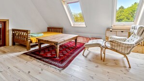 Un dormitorio tipo loft con techo inclinado, vigas de madera, una cama de madera, una mesa de comedor, un tapete rojo estampado, un sillón blanco y dos tragaluces.