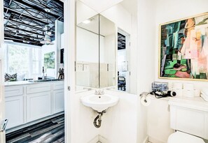 A modern bathroom with a white sink, a large mirror, and a painting above the toilet.