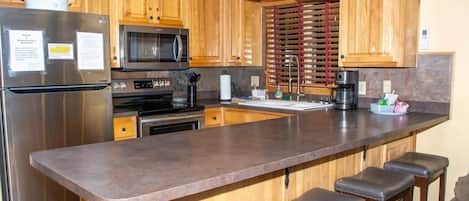 Una cocina con armarios de madera, un refrigerador de acero inoxidable, un microondas, un horno, una cafetera y una zona con taburetes de bar.