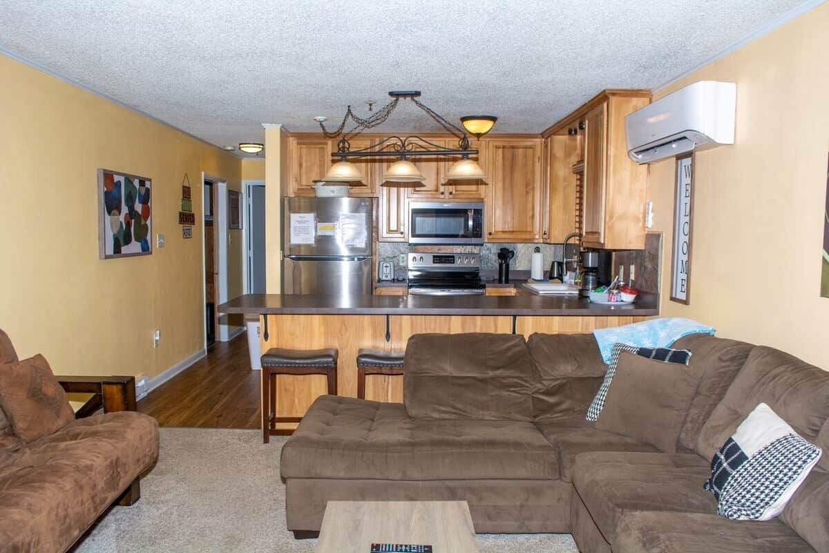 A living room with a brown sectional sofa, a coffee table, and a kitchen area with wooden cabinets, a refrigerator, and a microwave.