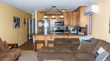 A living room with a brown sectional sofa, a coffee table, and a kitchen area with wooden cabinets, a refrigerator, and a microwave.
