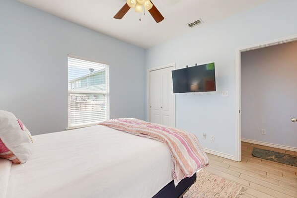 A bedroom with a bed, a ceiling fan, a TV mounted on the wall, and a window with blinds.
