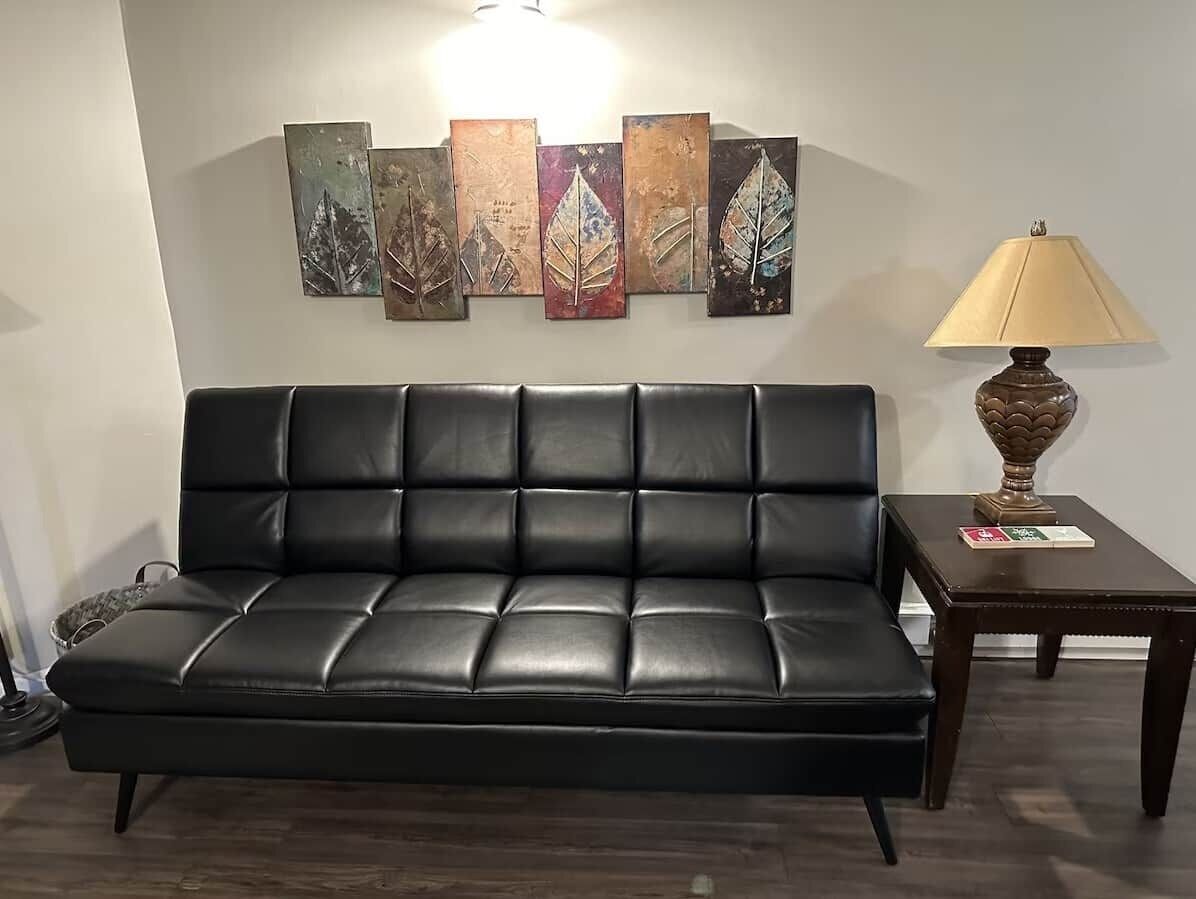Un canapé en cuir noir doté d’un dossier rembourré, placé contre un mur orné de plusieurs œuvres d’art aux motifs végétaux. Une petite table avec une lampe et un livre est posée sur le côté droit.