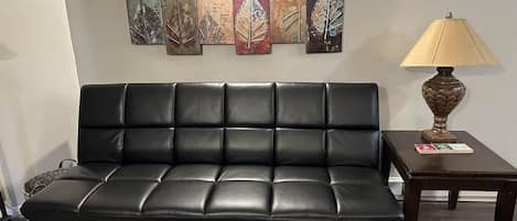 Un canapé en cuir noir doté d’un dossier rembourré, placé contre un mur orné de plusieurs œuvres d’art aux motifs végétaux. Une petite table avec une lampe et un livre est posée sur le côté droit.