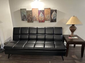 A black leather sofa with a cushioned backrest, positioned against a wall with a series of leaf-patterned artworks. A small table with a lamp and a book is placed to the right. - Winterplace D105 Ski in Ski out condo First Floor (Ghent)