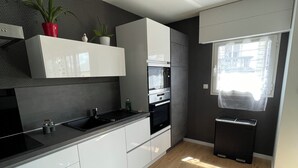 Una cocina moderna con armarios blancos, encimera negra y horno empotrado. Se ve una ventana con vistas al exterior y un pequeño cubo de basura.