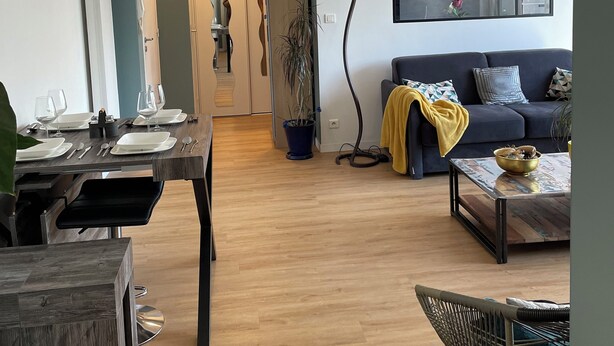 Una sala de estar moderna con un sofá oscuro, una mesa de centro y un comedor con una mesa dispuesta para dos personas.