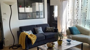 Una sala de estar moderna con un sofá azul oscuro, una mesa de centro con un cuenco, una lámpara de pie y un gran ventanal con cortinas.