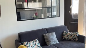 Una sala de estar moderna con un sofá azul oscuro, cojines estampados y una manta amarilla. Un espejo grande refleja el interior de la habitación, y se ve un ventilador de techo.