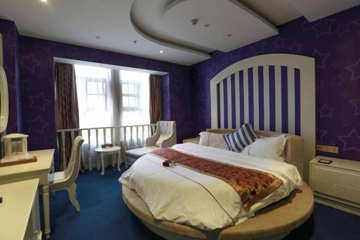 Hotelkamer met een rond bed, een klein bureau, een stoel, een nachtkastje en een raam met gordijnen.