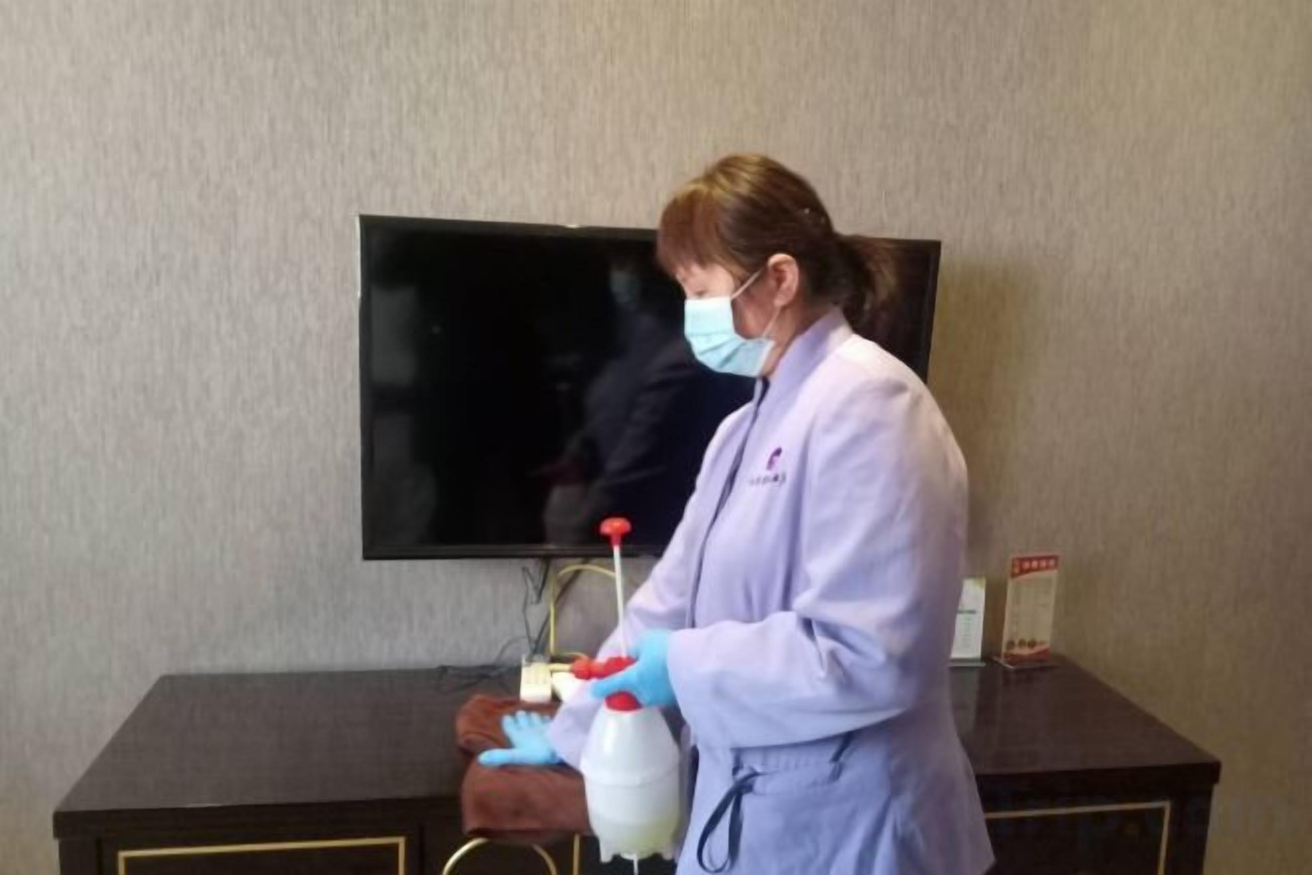 Una persona con bata de laboratorio y guantes de pie al lado de una mesa con una botella y un pulverizador.