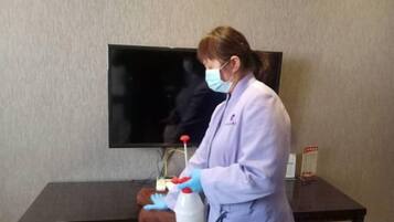 Una persona con bata de laboratorio y guantes de pie al lado de una mesa con una botella y un pulverizador.