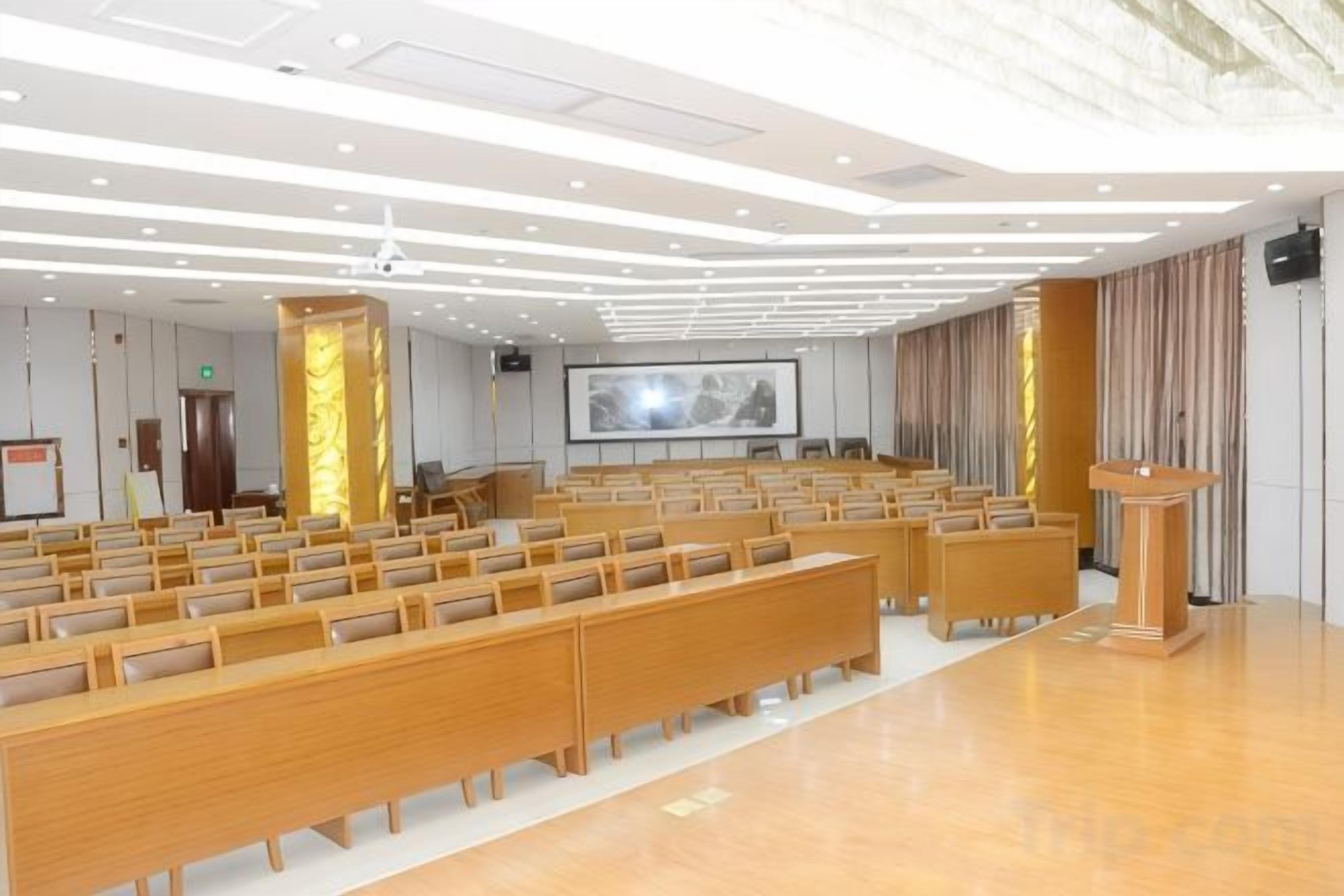 A conference room with rows of wooden chairs, a podium, and a large screen.
