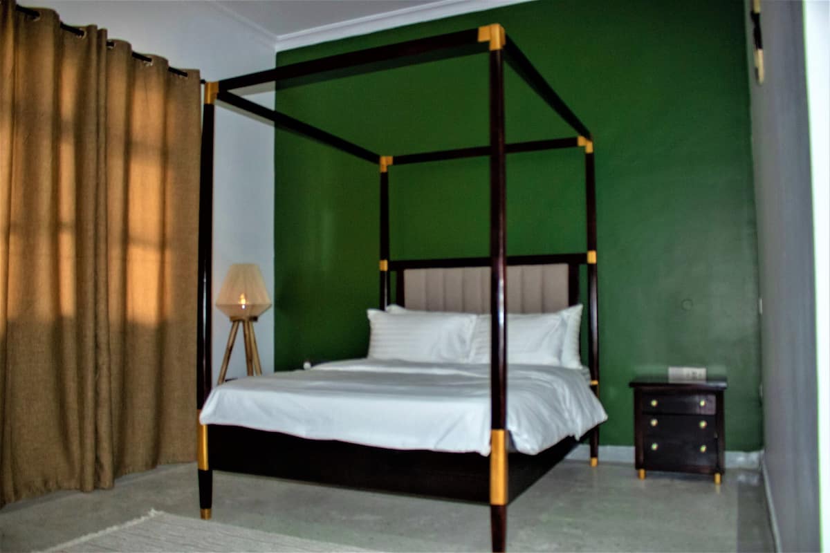 A four-poster bed with a wooden frame and white bedding in a room with green walls and a brown curtain.