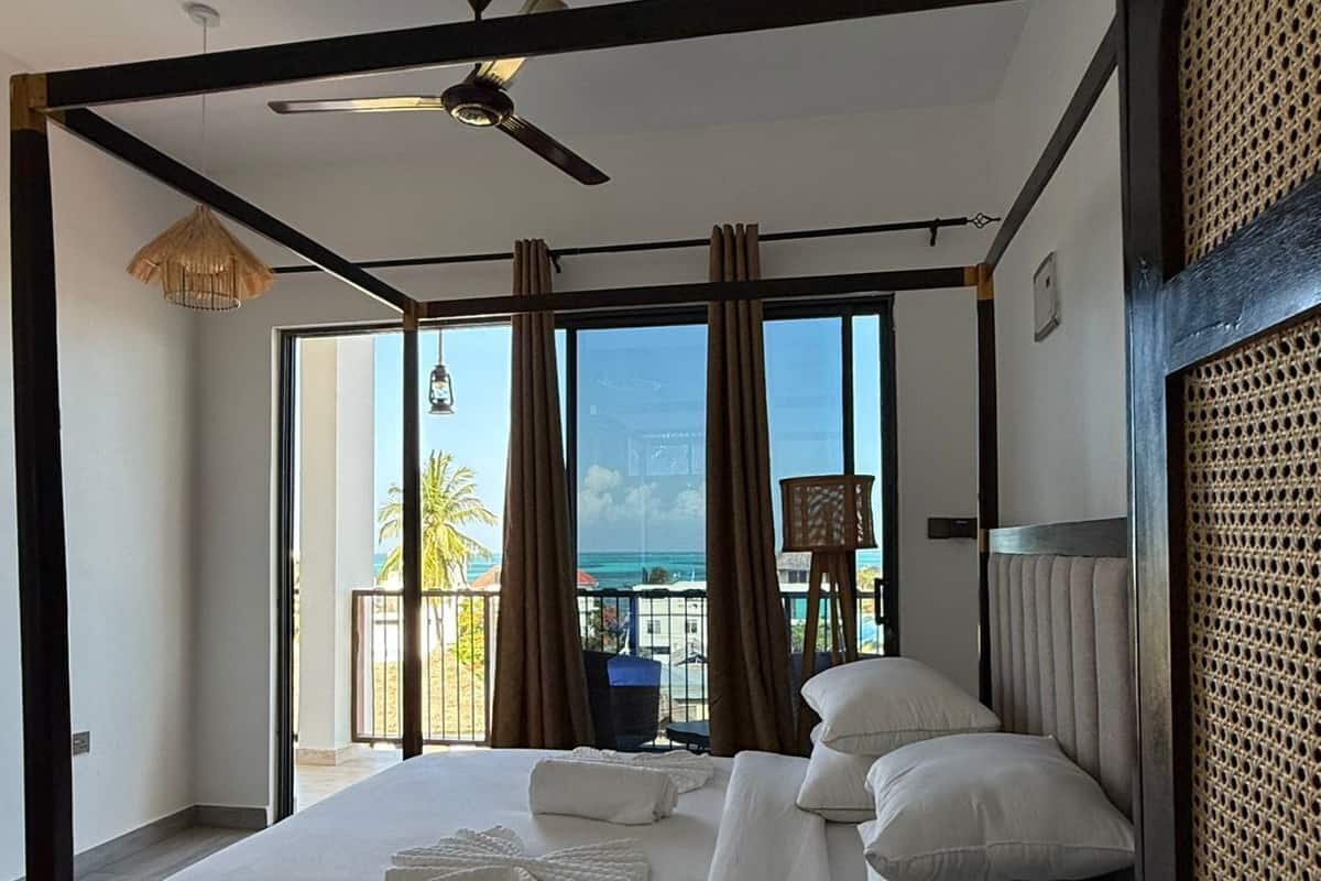 A four-poster bed with white linens, a ceiling fan, and a view of the ocean through large windows.
