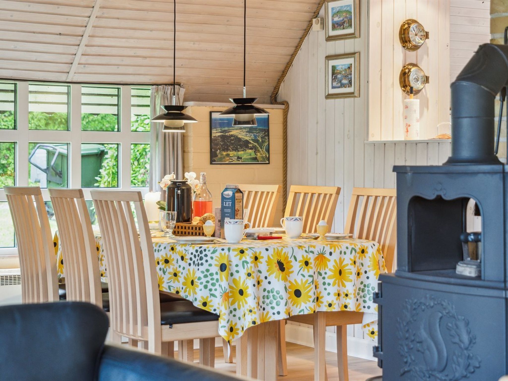 A dining area with a table set for four, surrounded by wooden chairs. A large stove is visible to the right, and there are framed pictures on the wall above the table.