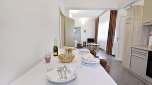 Un comedor con una mesa larga dispuesta para seis personas, una cocina al fondo y una sala de estar con televisor.