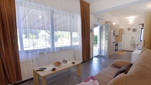 Un salón con sofá, una mesa de centro con controles remotos, una ventana con cortinas y una cocina al fondo.