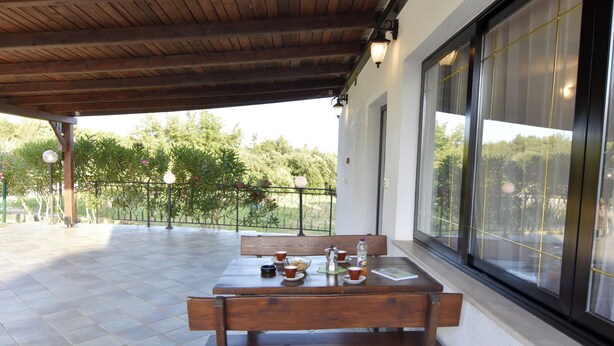 Un patio cubierto con una mesa y bancos de madera, piso de baldosas y vistas a la vegetación a través de amplias ventanas.
