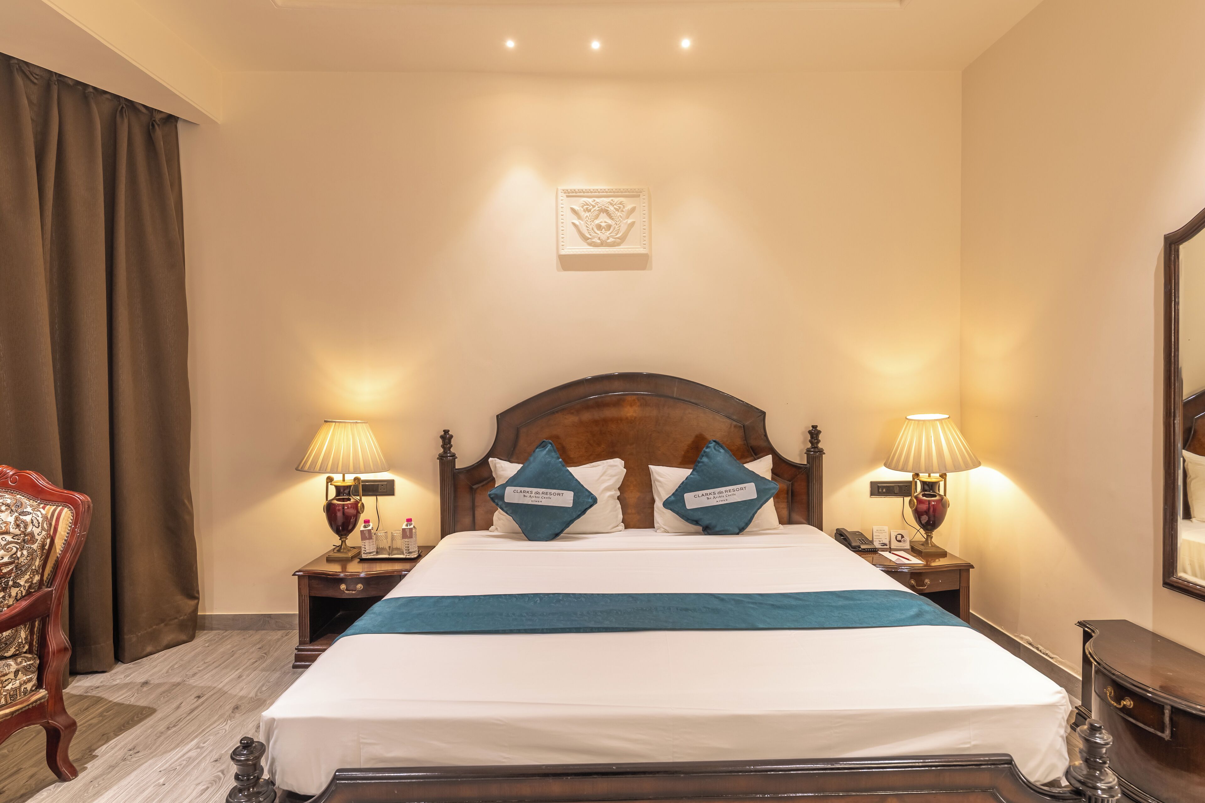 Een hotelkamer met een groot bed, twee nachtkastjes met lampen, een houten hoofdbord, een decoratief kussen en een nachtkastje met een fles, plus een stoel met een gestreept kussen.