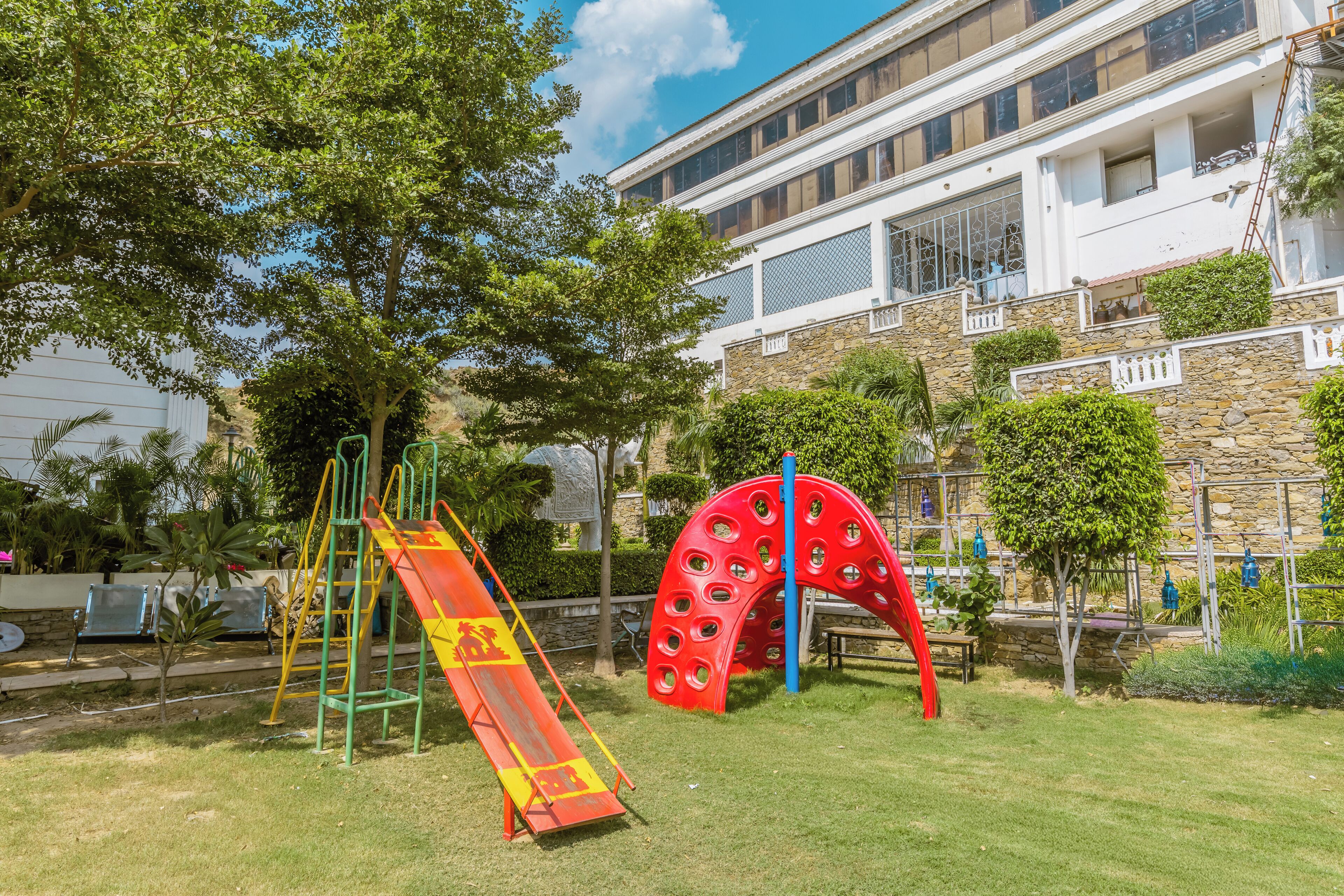 A playground with slides and climbing structures, surrounded by trees and a building with balconies.