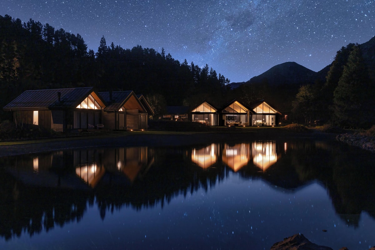 A lakeside cabin complex with multiple wooden cabins, each featuring large windows and a reflection in the calm water.