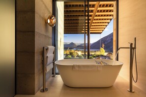 A modern bathroom with a freestanding bathtub, a glass shower enclosure, and a view of mountains and a lake.