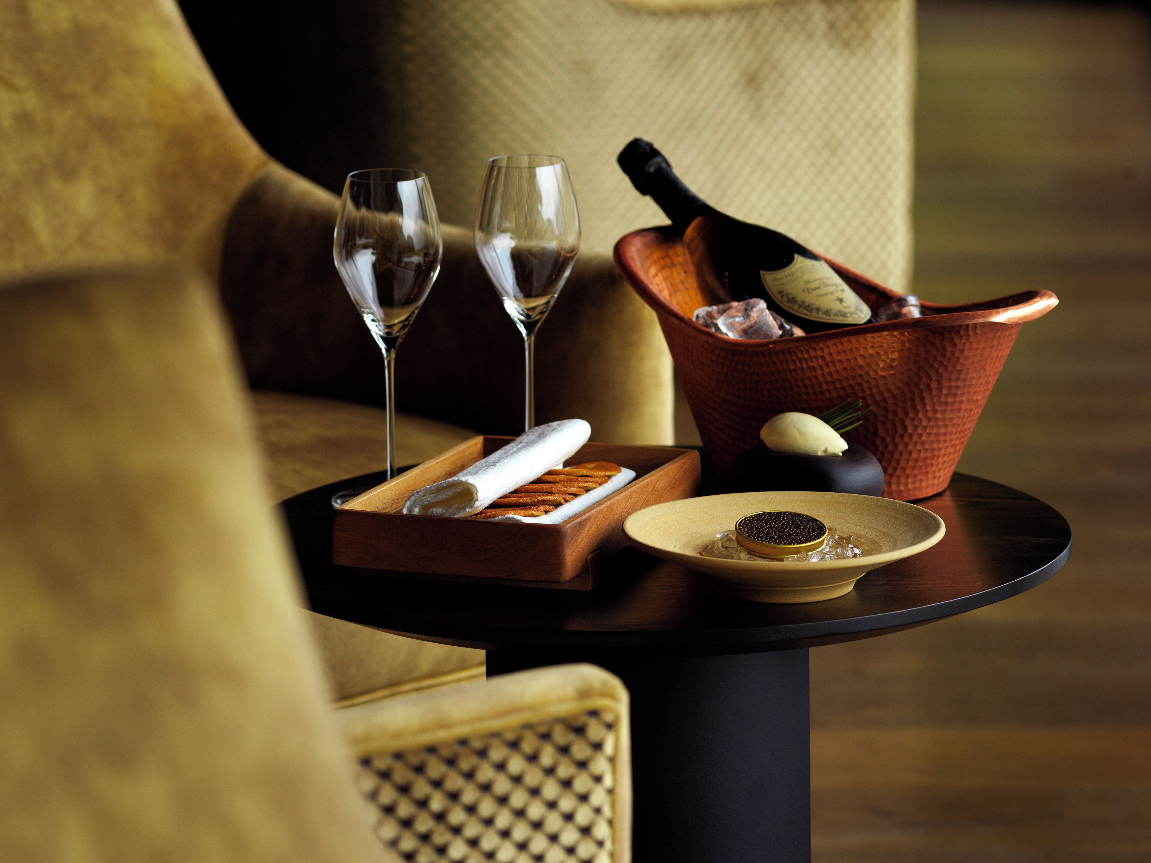A table with a bottle of champagne, two glasses, a plate with a dessert, and a tray with a napkin and a small bowl.