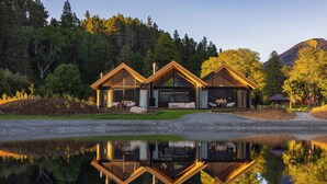A modern cabin with a glass facade, situated by a reflective lake, surrounded by forested hills. - Flockhill (Lake Pearson)