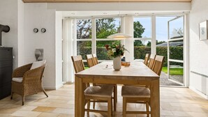 A dining area with a large table, chairs, and a vase of flowers. There are windows with a view of greenery outside.