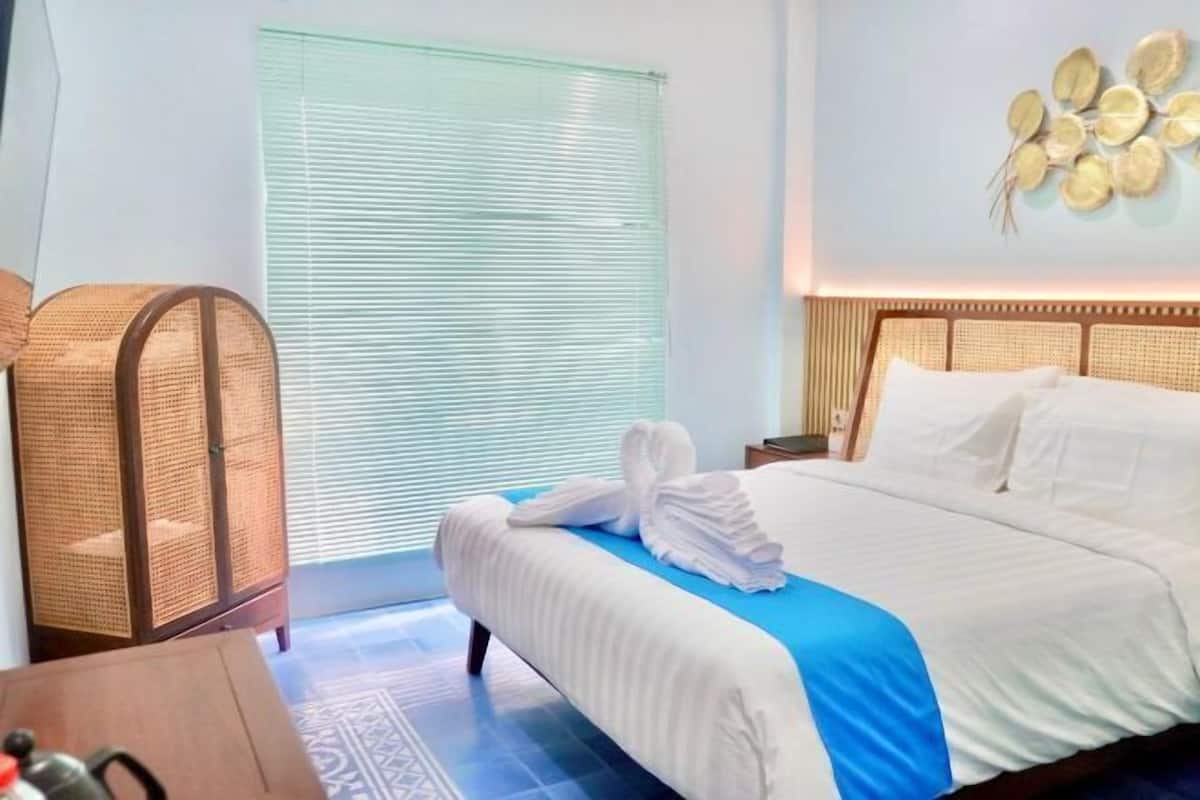Ein Hotelzimmer mit einem Bett, einem Korbsessel, einem Nachttisch mit Wasserkocher und einem Fenster mit Jalousien.