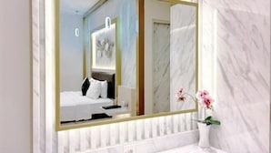 A modern hotel room with a large mirror, marble walls, a bed with white bedding, and a vase with flowers. - Grand Malioboro Yogyakarta Hotel (Gedong Tengen)
