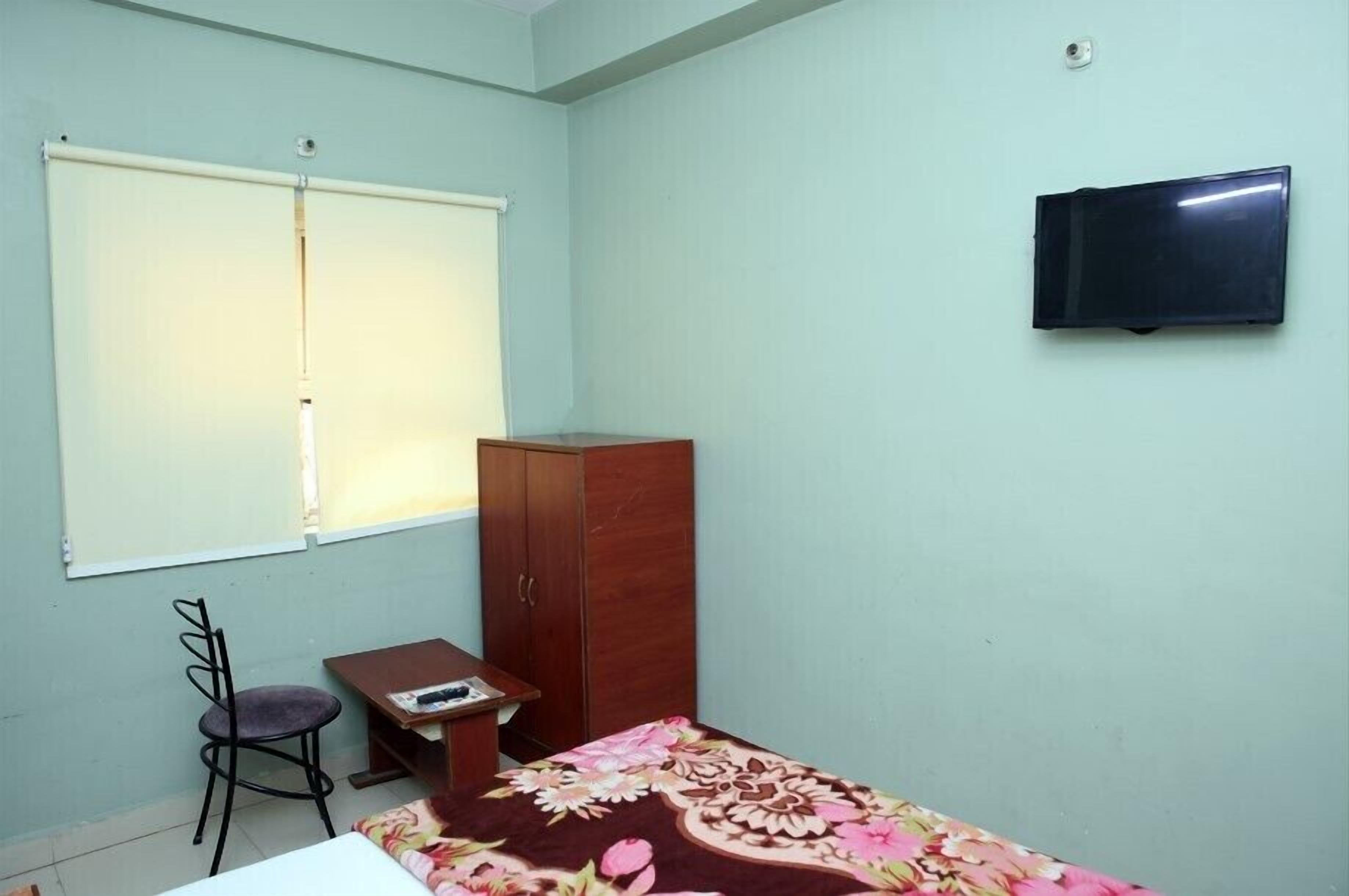Una habitación con cama, escritorio, silla, armario, ventana con persianas y televisor en la pared.