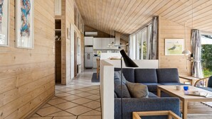 A modern interior with wooden walls and a tiled floor, featuring a sofa, a table, and a kitchen area with cabinets and a refrigerator.